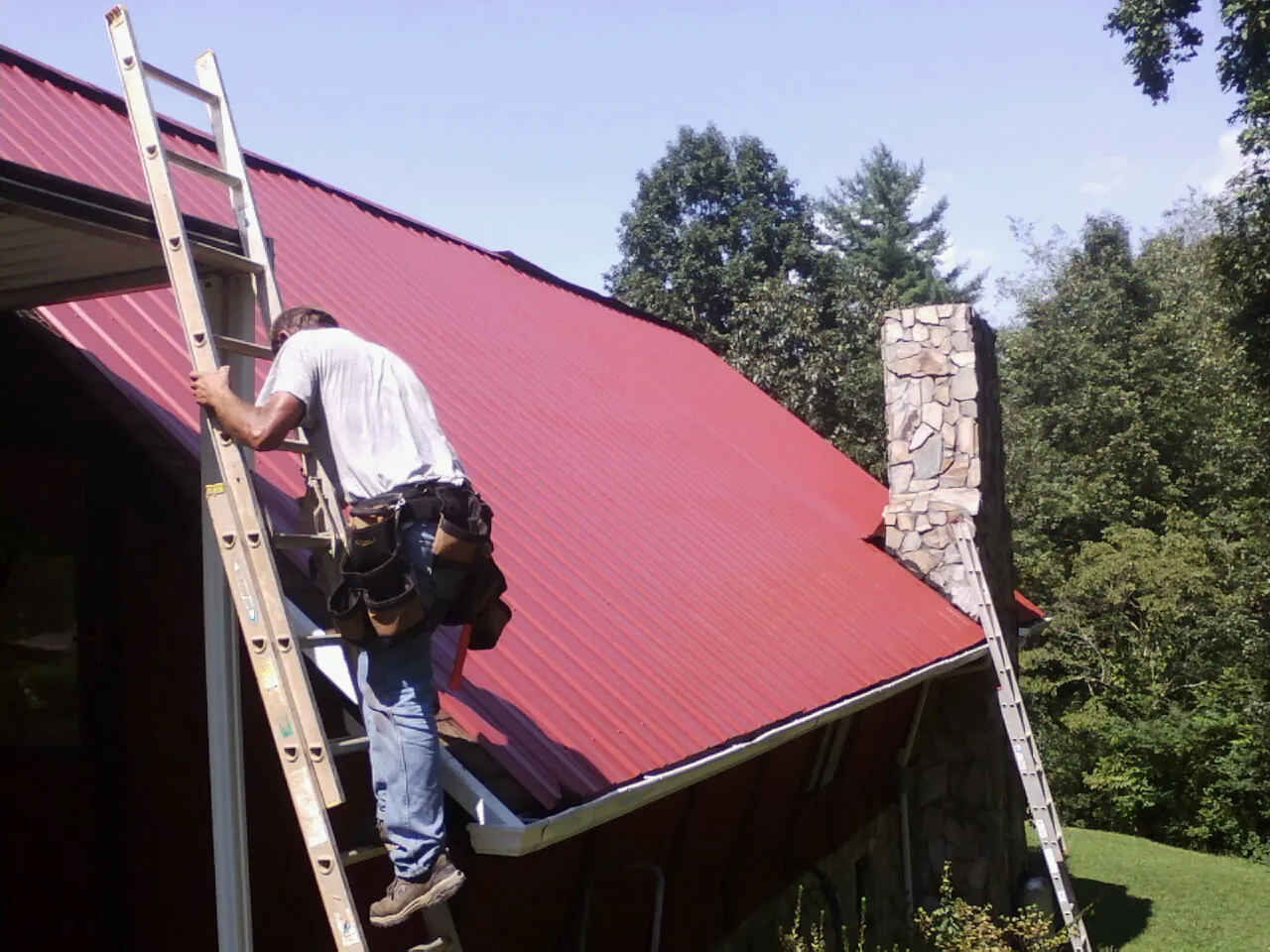 Roofer on a ladder beside a red metal roof and stone chimney — Soffit & Fascia Repair by Roofing Brothers in Highland Park